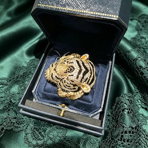 May include: A gold-toned tiger head brooch encrusted with clear crystals and accented with black enamel stripes. The brooch is displayed inside a dark navy blue, hinged jewelry box with a textured exterior and a soft, dark blue velvet interior lining.