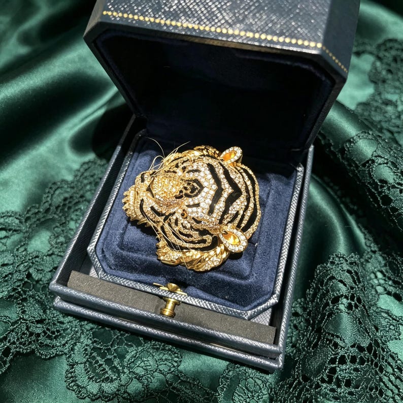 May include: A gold-toned tiger head brooch encrusted with clear crystals and accented with black enamel stripes. The brooch is displayed inside a dark navy blue, hinged jewelry box with a textured exterior and a soft, dark blue velvet interior lining.