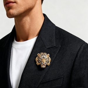 May include: A gold-toned tiger head brooch featuring intricate textured detailing, black enamel stripes, and small clear stones embedded across the face, pinned to the lapel of a black blazer worn over a plain white t-shirt.