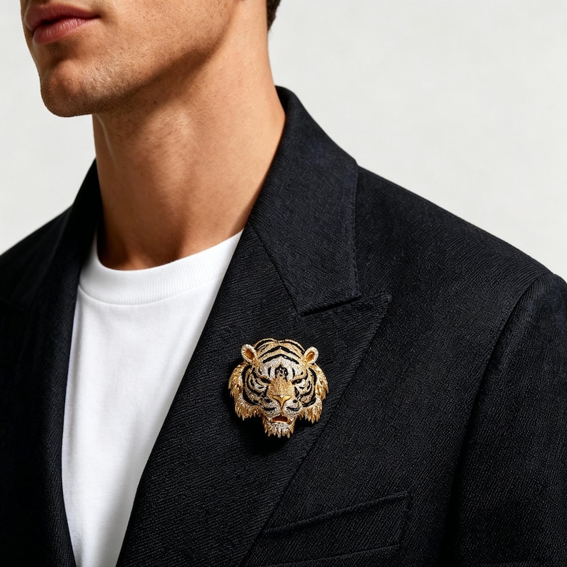 May include: A gold-toned tiger head brooch featuring intricate textured detailing, black enamel stripes, and small clear stones embedded across the face, pinned to the lapel of a black blazer worn over a plain white t-shirt.