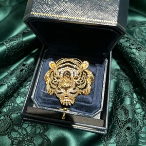 May include: A gold-toned tiger head brooch encrusted with clear crystals and black enamel stripes, featuring two small yellow crystal accents on the ears. The brooch is displayed inside a dark navy blue velvet-lined jewelry box with a textured lid.