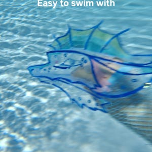 May include: A translucent blue and green aquatic creature toy with a finned back and tail, featuring dark blue outlines and the text "Easy to swim with" floating above it, submerged in clear, rippling water with visible light reflections.