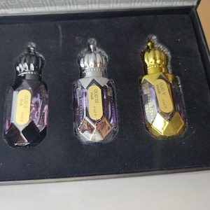 Pode incluir: Um conjunto de três frascos de perfume numa caixa de apresentação preta. Cada frasco tem uma tampa em forma de coroa e um corpo de vidro facetado com líquido roxo. Os rótulos dizem OUD II Musk 6 ml e, OUD III Musk 6 ml e e OUD V Malaki 6 ml e.
