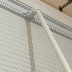 May include: A white roller shutter mechanism featuring a long, cylindrical operating rod attached to a plastic bracket. The shutter consists of horizontal slats that are closed, creating a uniform, textured surface across the window covering.