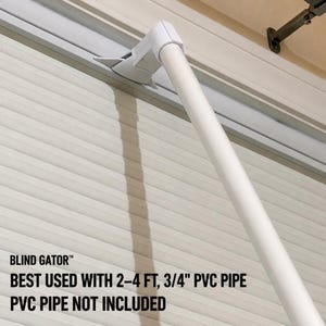 May include: A white plastic Blind Gator attachment connected to a horizontal window blind rail. The text reads: BLIND GATOR, BEST USED WITH 2-4 FT, 3/4" PVC PIPE, PVC PIPE NOT INCLUDED. The device is designed to hold a 3/4 inch PVC pipe.