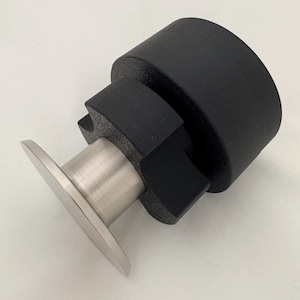 May include: A cylindrical black rubber door stop featuring a textured surface and a brushed stainless steel circular base plate. The object has a stepped design with a wider top section and a narrower middle section connected to the metal base.