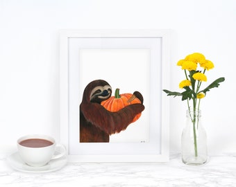 Puede incluir: Una impresión enmarcada en blanco de un perezoso marrón sosteniendo una calabaza naranja brillante. La impresión está sobre una superficie de mármol blanco con una taza de café y un jarrón de flores amarillas.