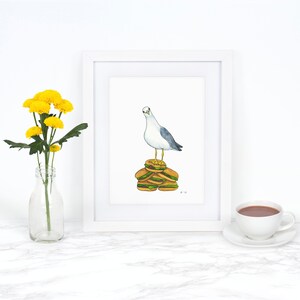 Puede incluir: Una pintura de acuarela de una gaviota de pie sobre una pila de cuatro hamburguesas. La gaviota est&aacute; mirando hacia la izquierda. La pintura est&aacute; enmarcada en un marco blanco y est&aacute; sobre una superficie blanca.