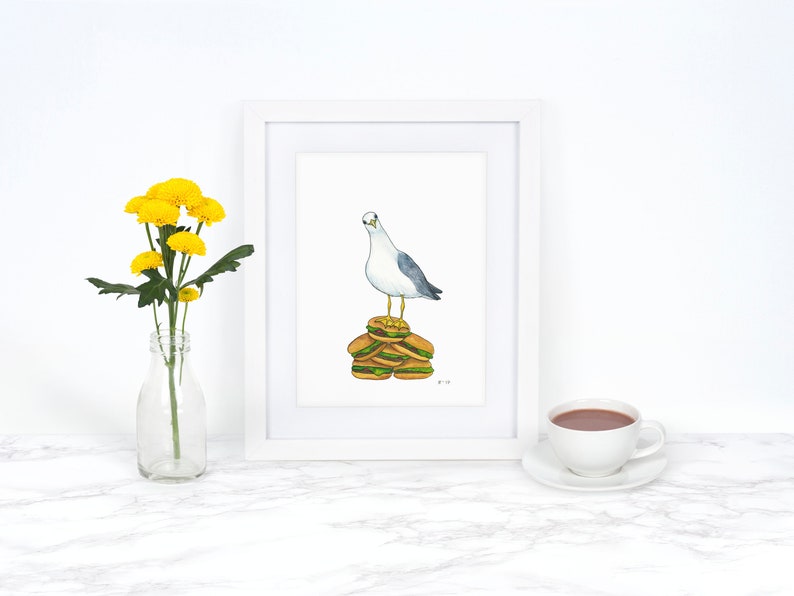 Puede incluir: Una pintura de acuarela de una gaviota de pie sobre una pila de cuatro hamburguesas. La gaviota est&aacute; mirando hacia la izquierda. La pintura est&aacute; enmarcada en un marco blanco y est&aacute; sobre una superficie blanca.