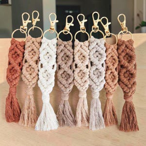 Puede incluir: Ocho llaveros de macramé en varios tonos de marrón, beige y crema, cada uno con un patrón de diamantes anudados, una borla inferior y un cierre de mosquetón de metal dorado unido a una anilla circular para una sujeción segura.