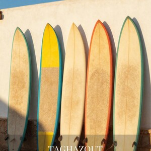 May include: Five surfboards lined up vertically, featuring varied colored rails in teal, yellow, orange, and green. The boards have textured, light-colored surfaces and small fins at the base. Text reads: TAGHAZOUT MOROCCO Surf Ocean Slow Life.