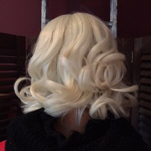 Puede incluir: Peluca rubia con capas suaves y rizadas. La peluca es de un color rubio claro y tiene un patr&oacute;n de ondas natural. La peluca est&aacute; peinada en un bob hasta los hombros con rizos. Se lleva una prenda negra debajo.