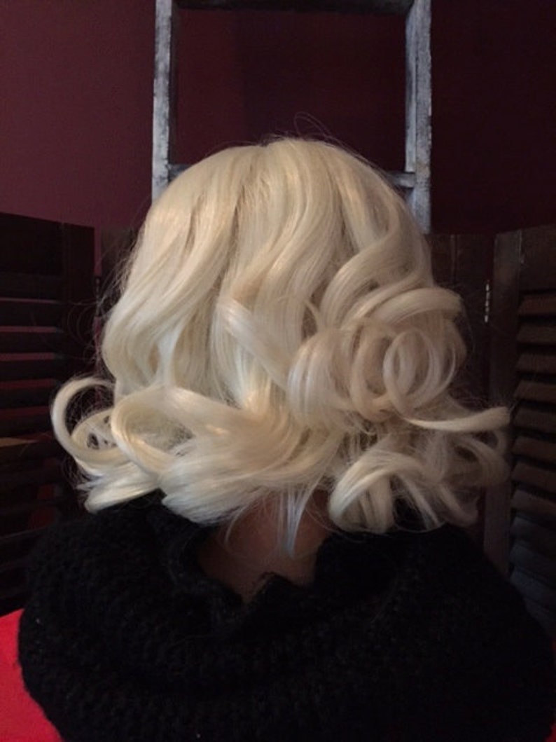 Puede incluir: Peluca rubia con capas suaves y rizadas. La peluca es de un color rubio claro y tiene un patr&oacute;n de ondas natural. La peluca est&aacute; peinada en un bob hasta los hombros con rizos. Se lleva una prenda negra debajo.