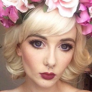 Puede incluir: Retrato en primer plano de una persona con cabello rubio y una corona floral de rosas rosas y blancas. La persona tiene maquillaje de ojos oscuro, un piercing en el tabique nasal y l&aacute;piz labial rojo oscuro.