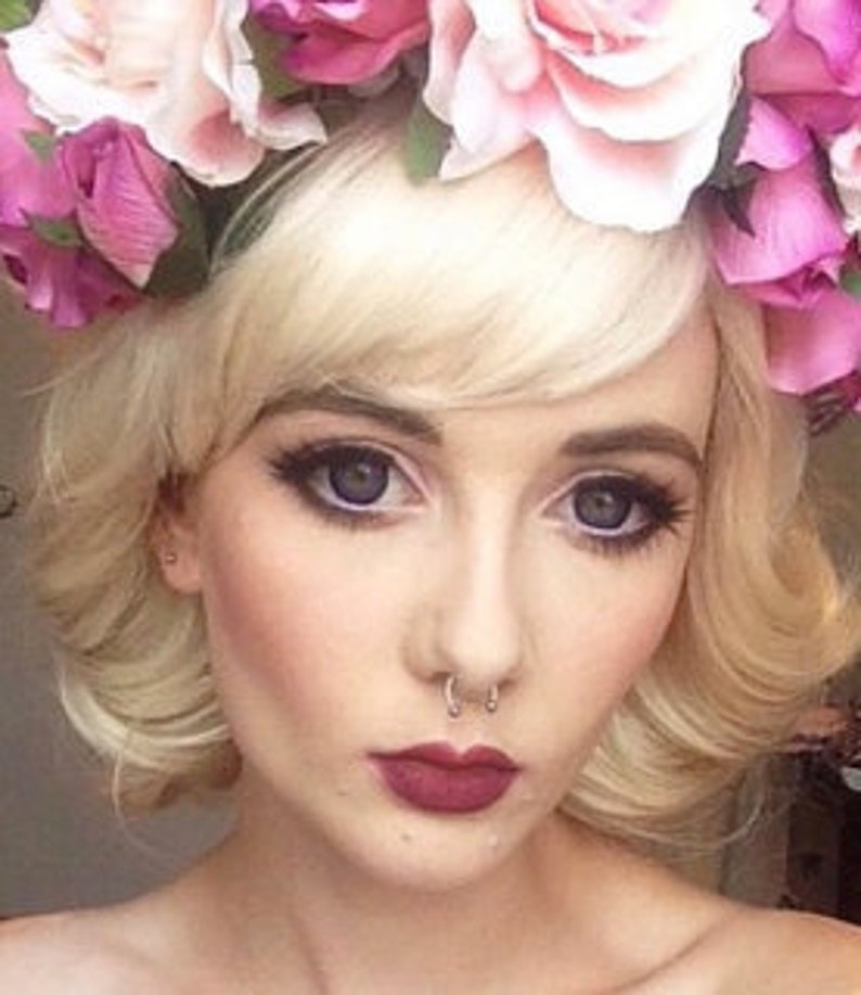 Puede incluir: Retrato en primer plano de una persona con cabello rubio y una corona floral de rosas rosas y blancas. La persona tiene maquillaje de ojos oscuro, un piercing en el tabique nasal y l&aacute;piz labial rojo oscuro.