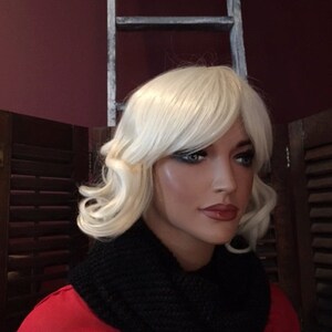 Puede incluir: Una cabeza de maniqu&iacute; con una peluca rubia y maquillaje de ojos oscuro. El maniqu&iacute; lleva un cuello de punto negro y una parte superior roja. Una escalera blanca desgastada est&aacute; en el fondo.