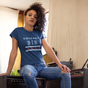 May include: A dark blue short-sleeve t-shirt featuring the text CHICAGO 312 in light blue, positioned above two light blue horizontal stripes and four red stars. The design is centered on the chest of the garment, which has a crew neckline.