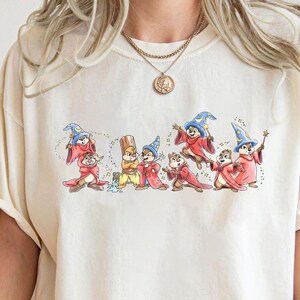Könnte beinhalten: Ein cremefarbenes Kurzarm-T-Shirt mit einem Grafikdruck von mehreren Zeichentrick-Streifenhörnchen, die als Zauberer in roten Roben und blauen spitzen Hüten gekleidet sind. Die Streifenhörnchen sind in verschiedenen Posen dargestellt, einige halten Zauberstäbe.