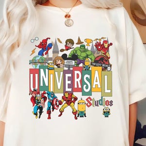 Könnte beinhalten: Ein cremefarbenes Kurzarm-T-Shirt mit einem bunten Grafikdruck verschiedener Charaktere und Logos. Der Text UNIVERSAL Studios ist mittig platziert und von Illustrationen von Superhelden, Zauberern und Minions umgeben.