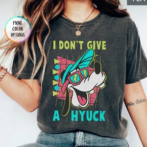 Könnte beinhalten: Ein anthrazitfarbenes Kurzarm-T-Shirt mit einer Grafik eines Zeichentrickhundes, der einen türkisfarbenen Hut und eine rosa Sonnenbrille trägt. Der Text I DON'T GIVE A HYUCK steht in lindgrünen und türkisfarbenen Buchstaben vor einem geometrischen Muster.