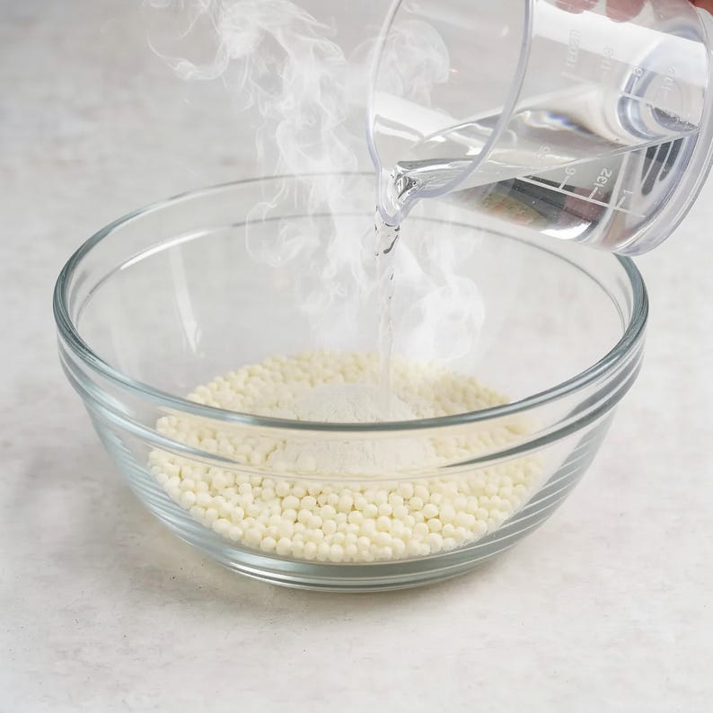 May include: A clear glass mixing bowl containing a layer of small, round, off-white wax beads. Hot water is being poured from a clear plastic measuring cup into the bowl, creating visible steam as it hits the beads and begins to melt them.