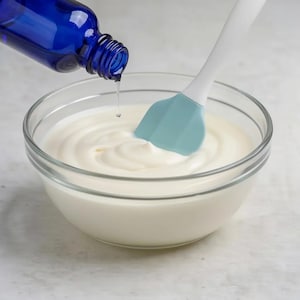 May include: A clear glass bowl filled with a smooth white cream, featuring a light blue silicone spatula resting inside. A dark blue glass dropper bottle is positioned above the bowl, dispensing a single clear liquid drop into the cream.