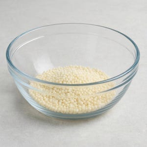 May include: A clear glass mixing bowl containing a mound of small, round, off-white wax beads. The bowl features a smooth, transparent surface with a slightly curved rim, and the beads are uniform in size, texture, and pale cream color.