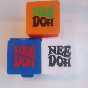 Op de afbeelding: Drie vierkante bakjes, één oranje, één blauw en één wit. Elk bakje is aan de voorzijde bedrukt met de tekst "NEE DOH". Het oranje bakje staat boven de blauwe en witte bakjes, die naast elkaar zijn geplaatst.