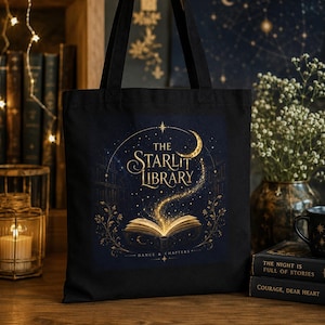 Könnte beinhalten: Eine schwarze Tragetasche mit einem Grafikdruck eines aufgeschlagenen Buches, aus dessen Seiten goldenes Licht strahlt. Der Text lautet THE STARLIT LIBRARY und DANCE & CHAPTERS. Das Design zeigt einen Halbmond, Sterne und florale Ranken.