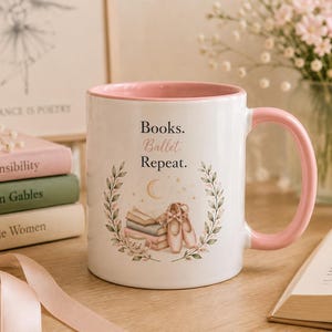 Könnte beinhalten: Ein weißer Keramikbecher mit rosafarbenem Innenraum und Henkel, verziert mit dem Text 'Books. Ballet. Repeat.' über einer Illustration von gestapelten Büchern, Ballettschuhen und einer Mondsichel, umrahmt von einem zarten grünen Blattkranz.