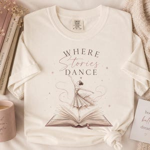 Könnte beinhalten: Ein cremefarbenes Kurzarm-T-Shirt mit einer Grafik einer Ballerina, die auf einem offenen Buch tanzt. Der Text "WHERE Stories DANCE" ist über der Illustration in einer Kombination aus Serifenschrift und Schreibschrift gedruckt.