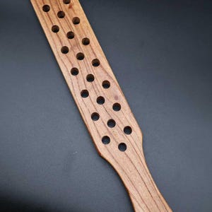 May include: A long, flat wooden paddle featuring a natural brown wood grain finish. The rectangular head of the paddle is perforated with two parallel rows of circular holes. A handle extends from the head, ending with a small hole and a black cord.