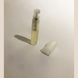 Könnte beinhalten: Ein kleines Parfüm-Probenfläschchen aus klarem Glas, gefüllt mit einer blassgelben Flüssigkeit, mit einem transparenten Kunststoffsprühkopf und einer separaten, passenden durchscheinenden Kunststoffkappe, die daneben auf einer hellen Fläche liegt.