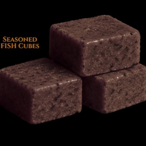 May include: Three dark brown, textured, square-shaped seasoned fish cubes stacked against a black background. The text "SEASONED FISH CUBES" is printed in orange to the left of the top cube. The surface of each cube appears dense and granular.
