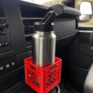May include: A stainless steel water bottle with a black lid sits inside a small, bright red plastic crate. The crate features a grid-like lattice design with integrated handles on the sides, providing a stable base for the bottle within a vehicle.