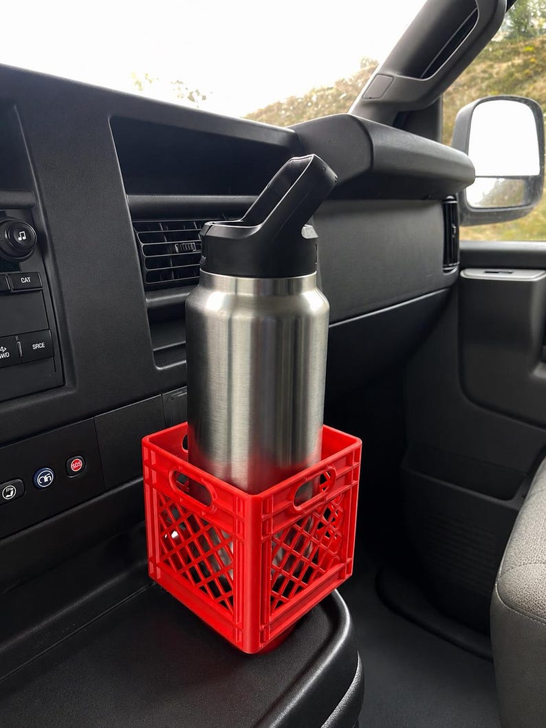 May include: A stainless steel water bottle with a black lid sits inside a small, bright red plastic crate. The crate features a grid-like lattice design with integrated handles on the sides, providing a stable base for the bottle within a vehicle.