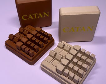 May include: Two sets of Catan game pieces in brown and beige. Each set includes a square storage box with the word CATAN printed in yellow on the front and a tray filled with small, molded plastic house and settlement game tokens.