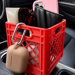 May include: A bright red plastic crate organizer designed for a vehicle cup holder. It contains a pink smartphone, a black quilted wallet, sunglasses, and a tube. A beige silicone case for wireless earbuds hangs from a silver carabiner on the side.