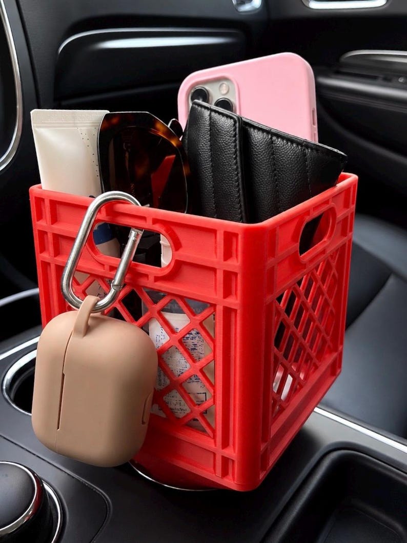 May include: A bright red plastic crate organizer designed for a vehicle cup holder. It contains a pink smartphone, a black quilted wallet, sunglasses, and a tube. A beige silicone case for wireless earbuds hangs from a silver carabiner on the side.