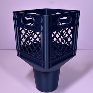 May include: A dark blue plastic cup holder insert designed to fit into a vehicle console. The top section features a square, open-grid crate pattern with integrated handles on each side, transitioning into a solid, tapered cylindrical base.