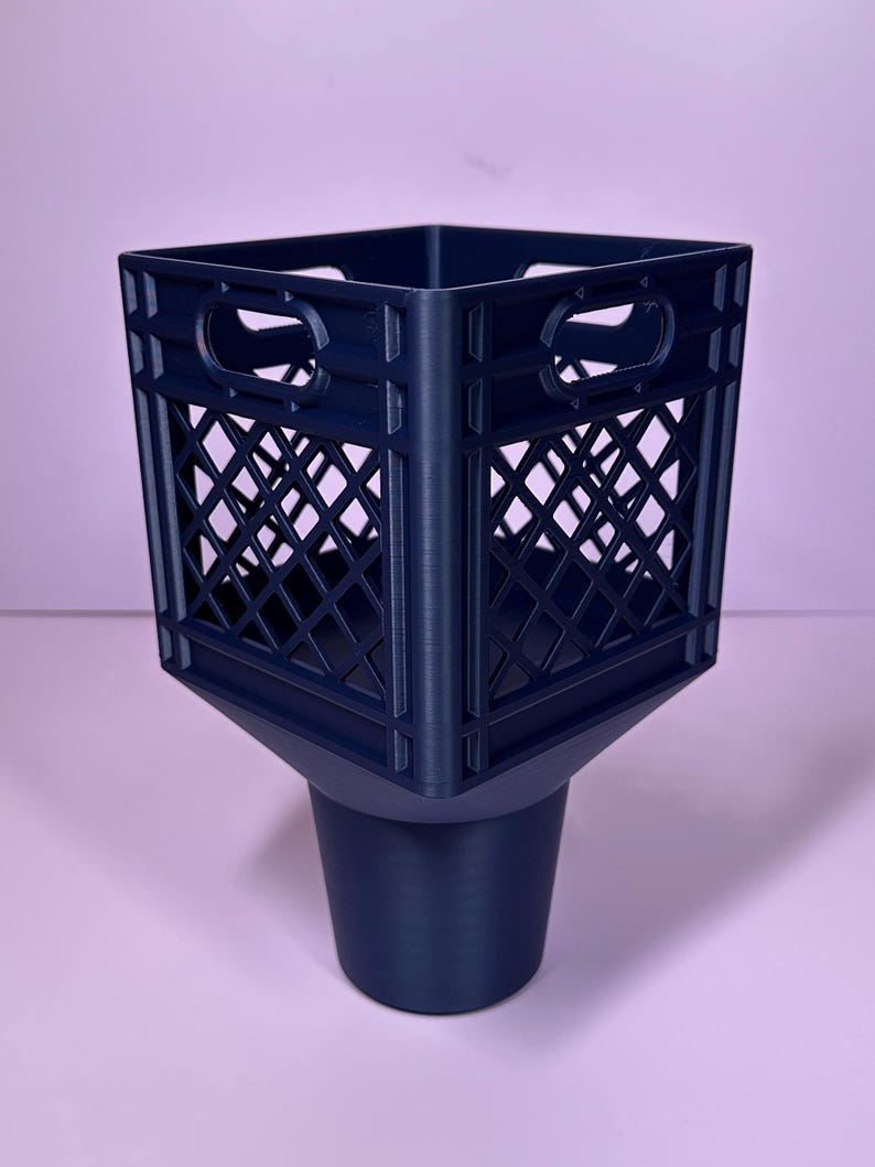 May include: A dark blue plastic cup holder insert designed to fit into a vehicle console. The top section features a square, open-grid crate pattern with integrated handles on each side, transitioning into a solid, tapered cylindrical base.