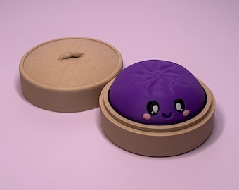 May include: A purple, rounded, smiling character with large black and white eyes and pink cheeks, nestled inside a tan, circular, lidded container that resembles a bamboo steamer. The lid is placed to the left of the base.
