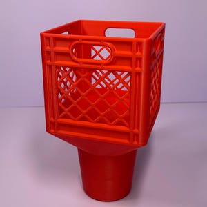 May include: A bright orange plastic cup holder featuring a square, crate-style top section with a lattice pattern and integrated handles, transitioning into a solid, tapered cylindrical base designed to hold a beverage container securely.
