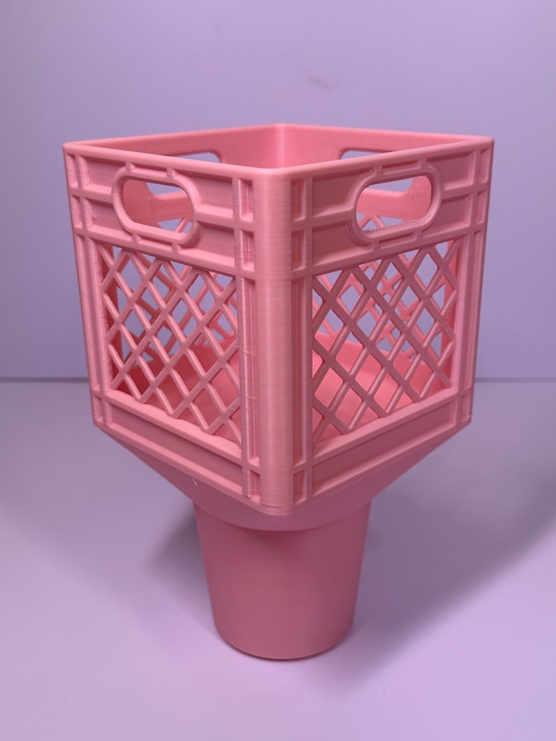 May include: A light pink plastic storage container featuring a square, open-grid crate top with integrated handles on each side, seamlessly connected to a solid, tapered cylindrical base, creating a singular, unified desk organizer structure.