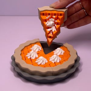 May include: A 3D printed pumpkin pie toy with a gray pie tin, a tan crust, orange filling, and white dollops of whipped cream. One triangular slice is detached from the main pie, revealing the segmented, blocky construction of the object.