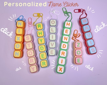 May include: A collection of six personalized name clicker keychains in various colors, each featuring square buttons with letters spelling out names including Nixon, Story, Rhyder, Olive, Hendrix, Zoe, and Mavis, attached to colorful plastic clips.