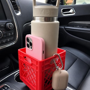 May include: A bright red plastic crate-style cup holder containing a beige YETI water bottle with a handle and a light pink smartphone. A beige wireless earbud case is attached to the side of the red crate with a silver metal carabiner clip.