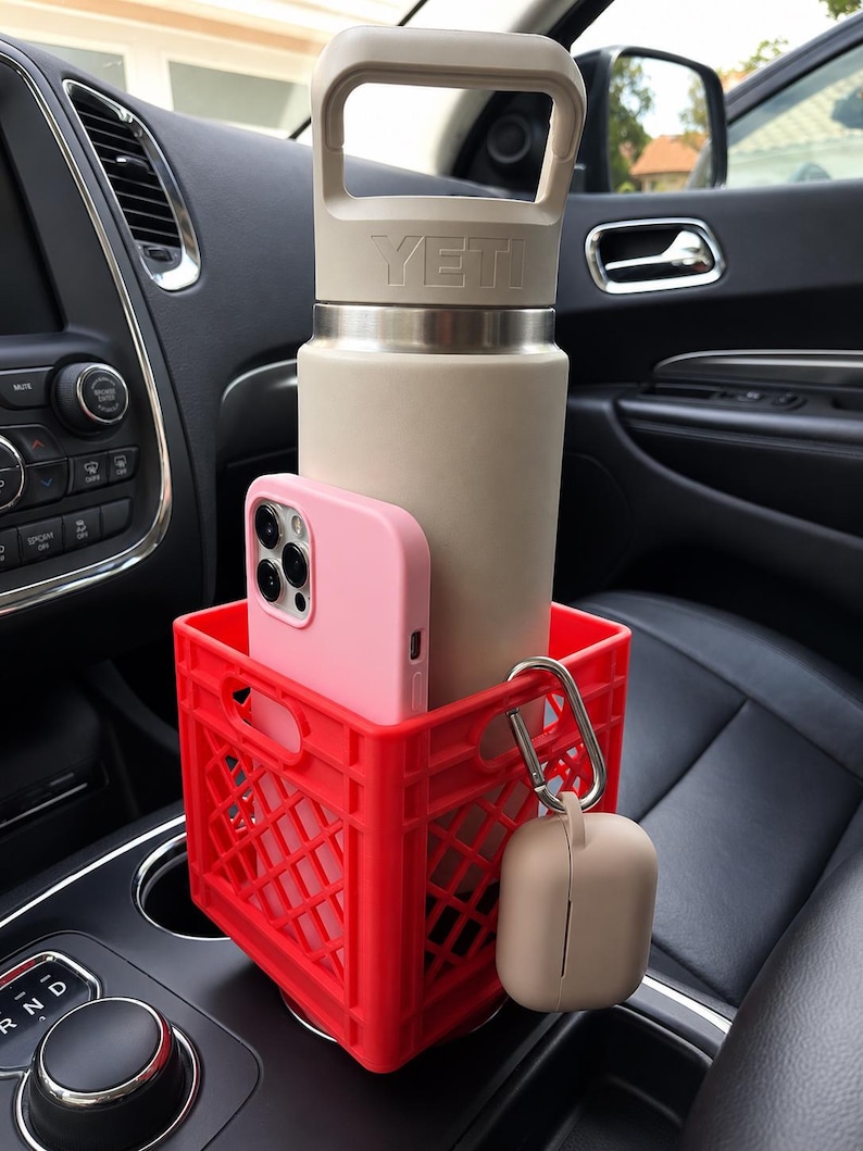 May include: A bright red plastic crate-style cup holder containing a beige YETI water bottle with a handle and a light pink smartphone. A beige wireless earbud case is attached to the side of the red crate with a silver metal carabiner clip.