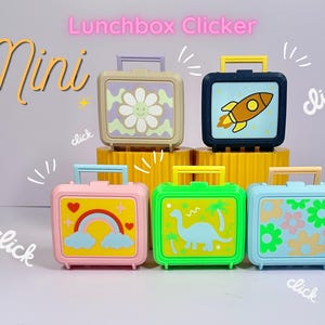 May include: Five small plastic lunchbox clicker toys with handles. The designs include a daisy, a rocket ship, a rainbow with hearts, a dinosaur with palm trees, and a floral pattern. The text "Mini" and "Lunchbox Clicker" appears above the toys.
