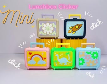 May include: Five small plastic lunchbox clicker toys with handles. The designs include a daisy, a rocket ship, a rainbow with hearts, a dinosaur with palm trees, and a floral pattern. The text "Mini" and "Lunchbox Clicker" appears above the toys.